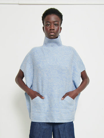 A person with short hair is wearing a light blue, oversized Square Poncho from Zero + Maria Cornejo made from sustainably-sourced cashmere, featuring short sleeves and pockets. Paired with dark blue pants, they stand against a plain white background, looking straight at the camera with a neutral expression.