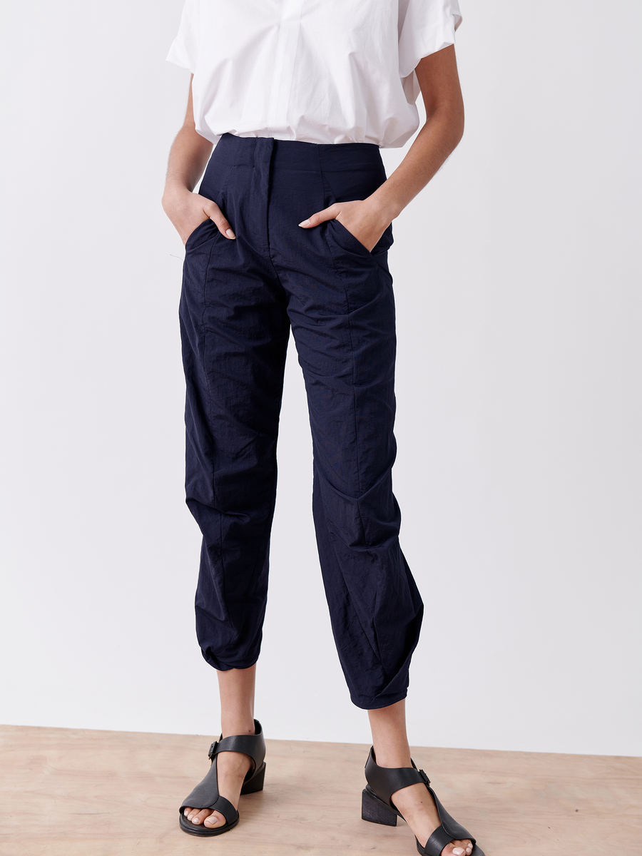 A person wears a white short-sleeve shirt tucked into high-waisted Takeo Pant by Zero + Maria Cornejo, featuring a crinkled texture in navy tech taffeta, hands in pockets. Black open-toe block-heeled sandals complete the sleek, modern look against a light background.