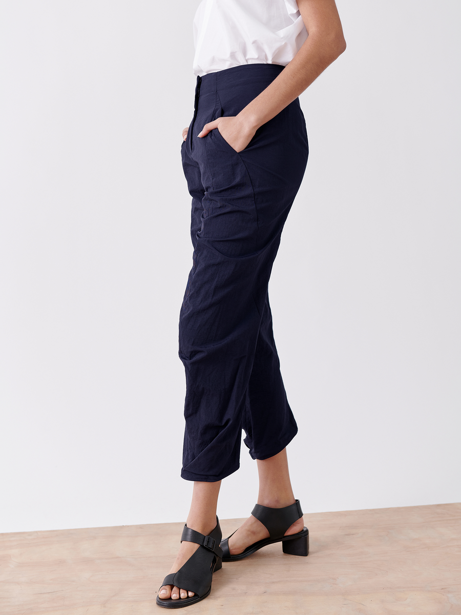 A person is wearing the Takeo Pant by Zero + Maria Cornejo, a navy blue tech taffeta cropped pant, with one hand in the pocket. They pair it with a white shirt and black chunky-heeled sandals against a plain white background.