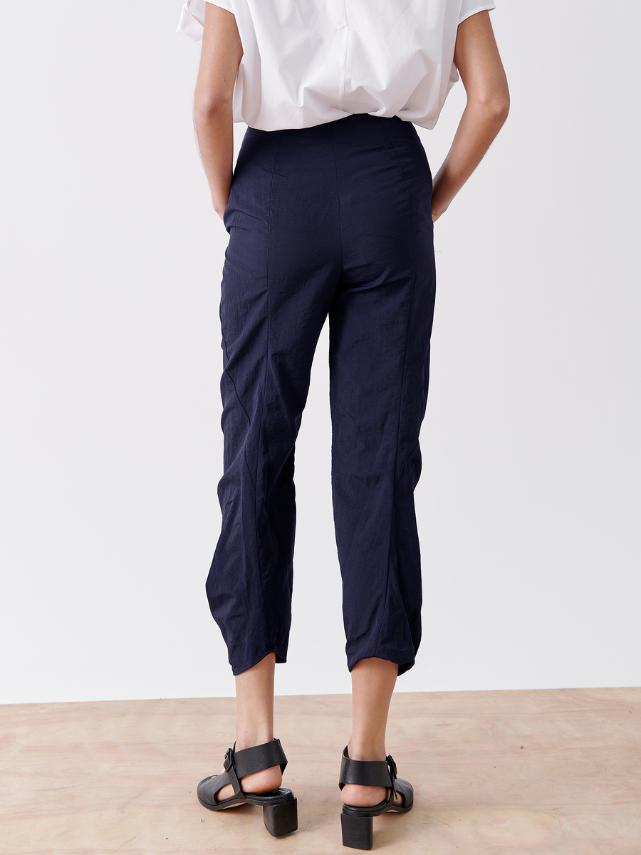 A person stands on a wooden floor, wearing a white shirt and Zero + Maria Cornejo's Takeo Pant in navy blue tech taffeta. Their hands rest in pockets, while black strappy sandals complete the look. The water-repellent fabric ensures packable weight for travel against the white backdrop.