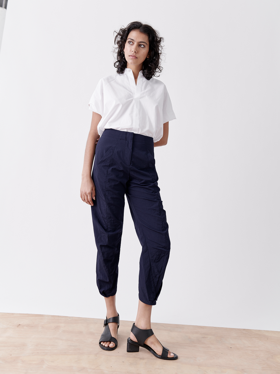 A person with curly hair stands on a wooden floor against a white backdrop. They're wearing a white short-sleeved shirt, Zero + Maria Cornejo's dark blue Takeo Pant made from water-repellent tech taffeta, and black heeled sandals. Relaxed and looking aside, they embody packable weight for travel.