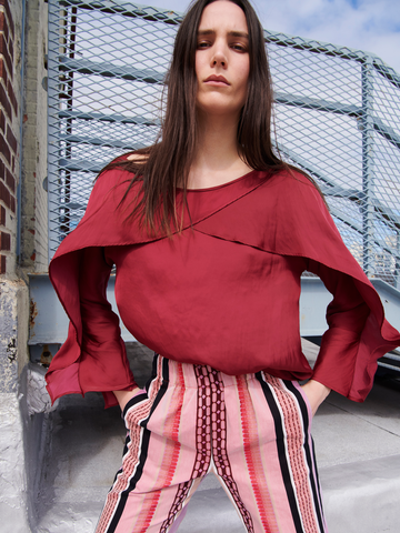 A woman with long brown hair wears the Ido Shirt by Zero + Maria Cornejo, featuring fluttery long sleeves, paired with colorful striped pants. She stands confidently against an industrial metal backdrop on a rooftop under a partly cloudy sky, enhancing the vibrant New York vibe.