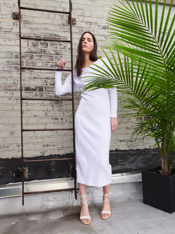 A woman in a Rib Long-Sleeved Ama Dress by Zero + Maria Cornejo and strappy heels leans against a metal ladder on a brick wall, with a large potted palm enhancing the minimalist urban setting.