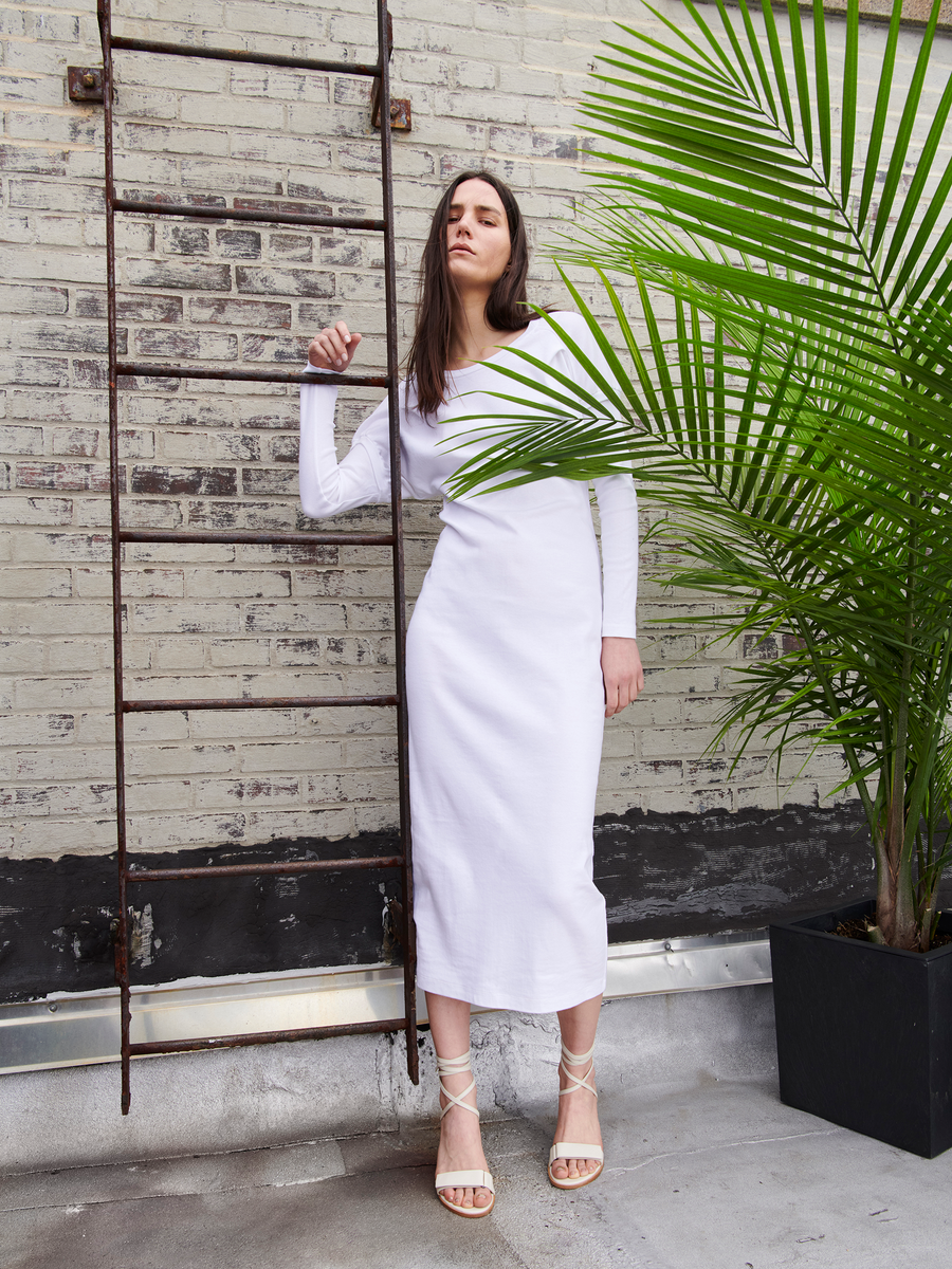 A woman in a Rib Long-Sleeved Ama Dress by Zero + Maria Cornejo and strappy heels leans against a metal ladder on a brick wall, with a large potted palm enhancing the minimalist urban setting.