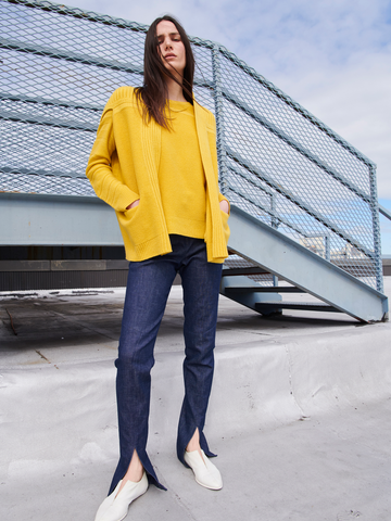 A person standing on a rooftop near a metal railing, hands in their pockets, wears Zero + Maria Cornejo's bright yellow organic cotton sweater, dark blue Denim Tala Trouser with hem slits, and white shoes. The sky above is partly cloudy.