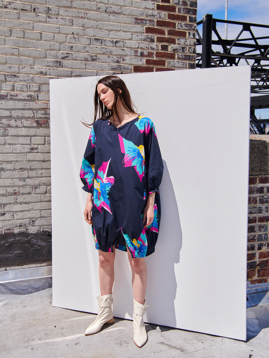 A person stands outdoors against a white backdrop, wearing the Long-Sleeved Luca Dress by Zero + Maria Cornejo, featuring a dark silhouette with bright abstract floral patterns, paired with white ankle boots. The setting includes a brick wall and metal scaffolding under a clear sky.