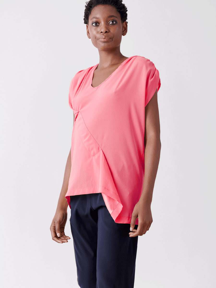 A person in a bright pink Ruched Fold Top by Zero + Maria Cornejo, styled with an asymmetrical front, pairs it with dark pants. They stand smiling against a plain white background.