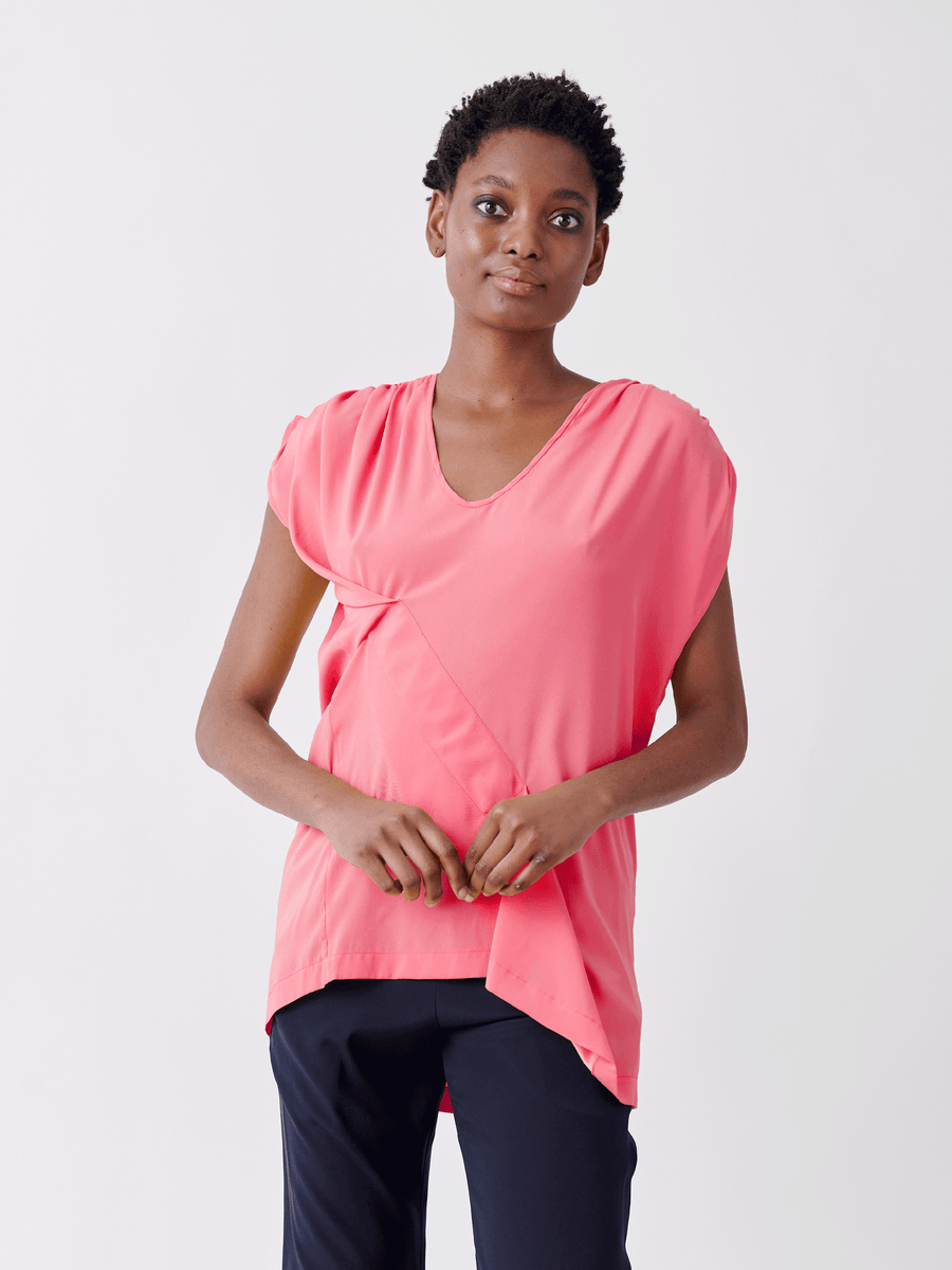 A person in a stylish pink Ruched Fold Top by Zero + Maria Cornejo, paired with dark pants, stands against a white backdrop, calmly clasping their hands.