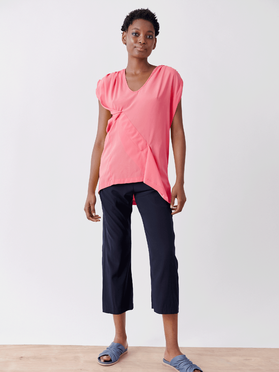 A person stands against a plain background, wearing the Ruched Fold Top by Zero + Maria Cornejo in pink silk charmeuse, black cropped pants, and blue slide sandals. Their hands are relaxed by their sides.