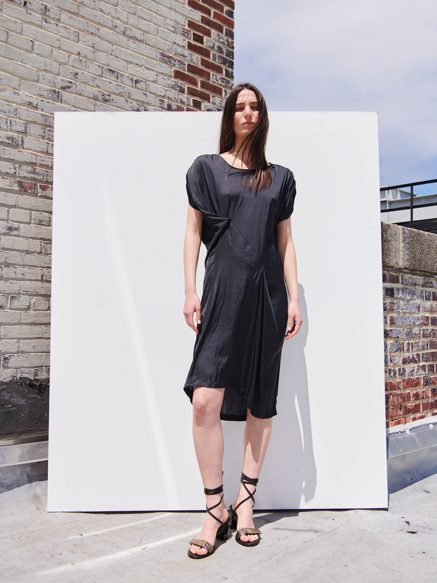 A woman in a Ruched Fold Dress by Zero + Maria Cornejo and strappy sandals stands before a whiteboard on a brick-walled rooftop, her long brown hair cascading under a clear sky.