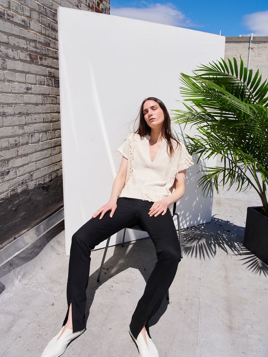 A person with long hair sits on a chair outdoors with a potted plant beside them, wearing a cream blouse, Denim Tala Trouser by Zero + Maria Cornejo, and white shoes against a white backdrop on a rooftop under clear skies.