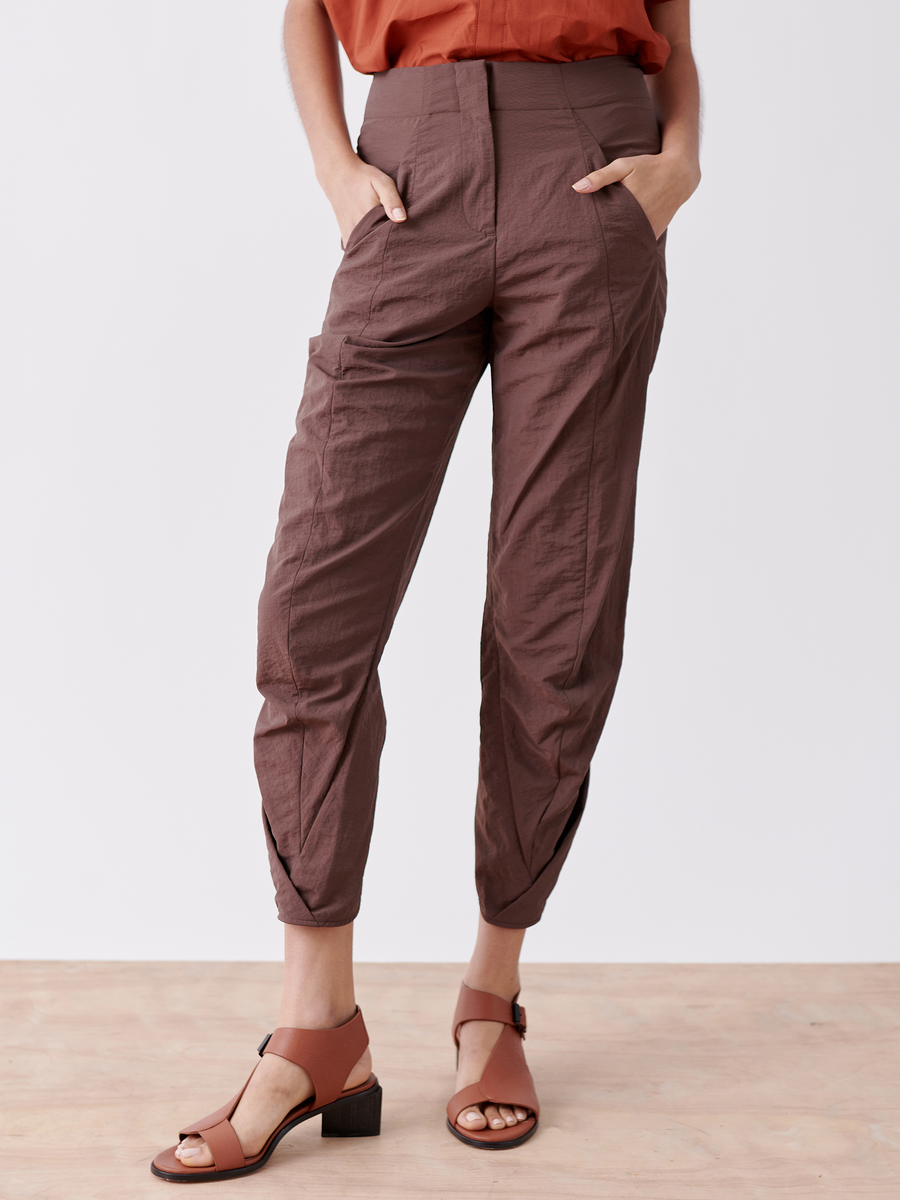 Someone wearing the Zero + Maria Cornejo Takeo Pant - Final Sale in brown, textured tech taffeta with hands in pockets, paired with tan heeled sandals and a warm rust upper garment stands out against a plain white background. The lightweight design is practical for travel.