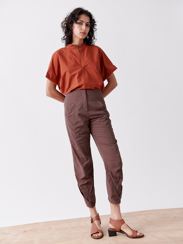 A person with curly hair wears a rust-colored, short-sleeved blouse and Zero + Maria Cornejo's Takeo Pant. They stand on a wooden floor in brown sandals, facing the camera against a plain white background—ideal for packable adventures.