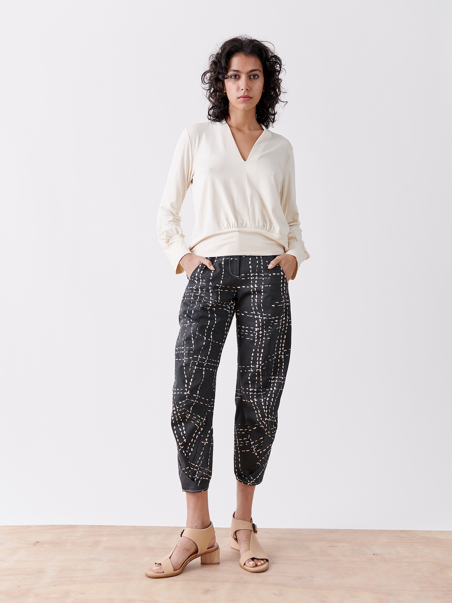 Against a white background, a person with curly hair wears a cream organic cotton top paired with Zero + Maria Cornejo's Takeo Pant in black printed denim featuring an abstract white pattern. They complete the look with beige open-toe sandals, hands casually in their pockets.