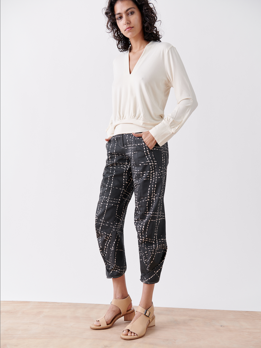 A person with curly hair stands against a white background wearing a cream-colored organic cotton long-sleeve top and the black printed Takeo Pant from Zero + Maria Cornejo. They complete the look with beige open-toe sandals.