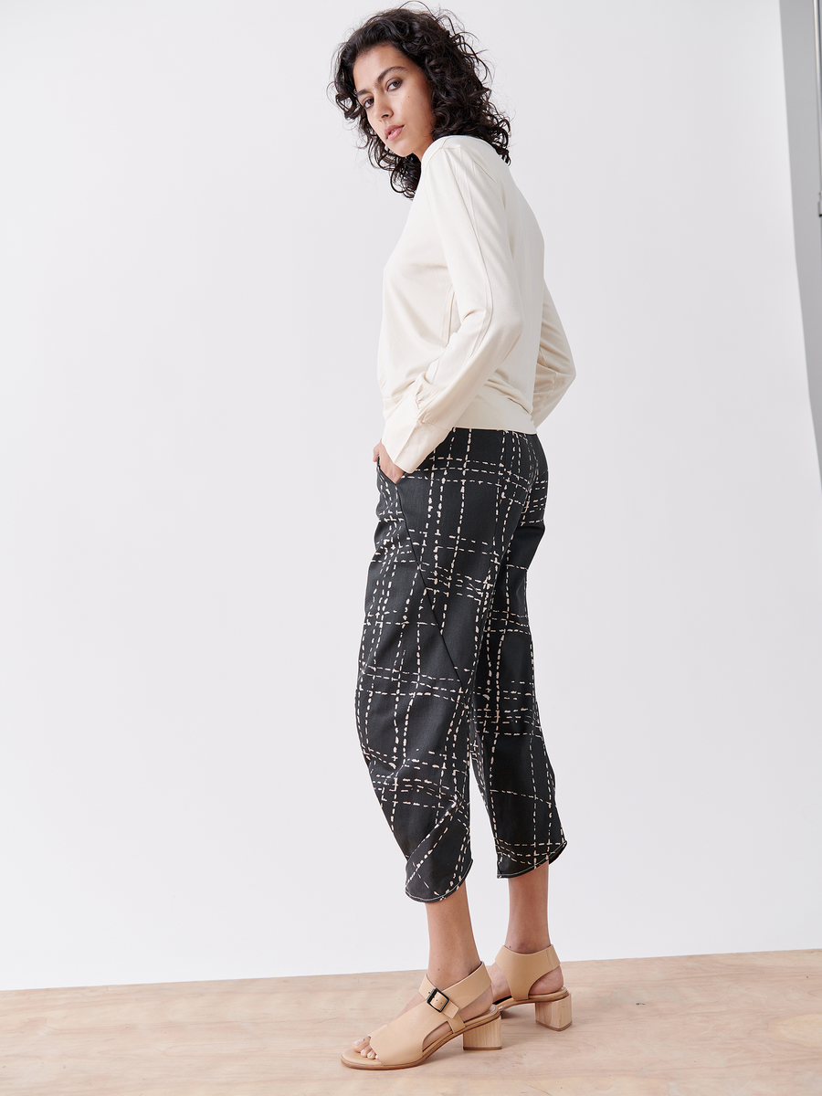 Wearing the Zero + Maria Cornejo Takeo Pant, a person stands on a wooden floor against a white backdrop. They're in a black printed denim pant and cream long-sleeve top, paired with light-colored sandals, striking a relaxed pose while looking off to the side.