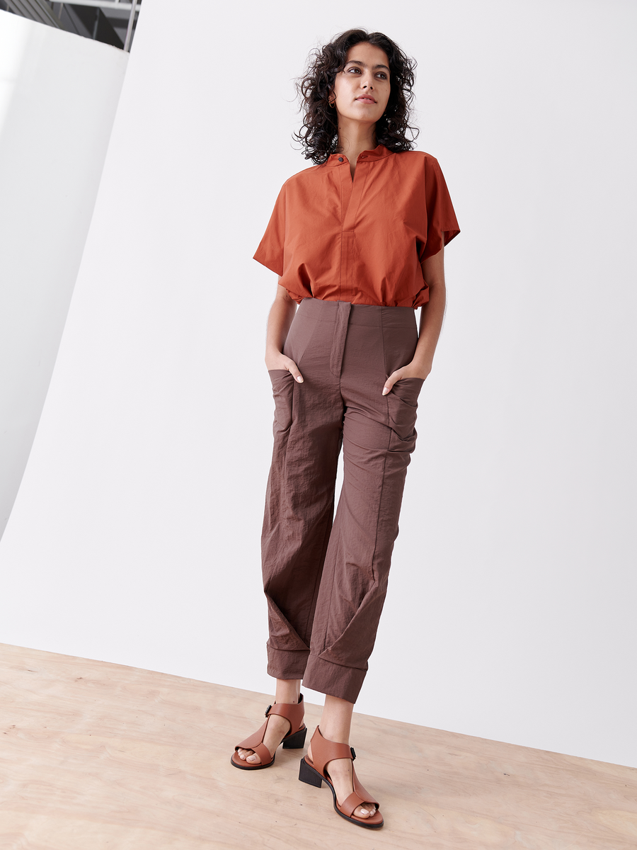 A woman with curly hair stands confidently in a minimalist room, wearing the Akeo Jodhpur by Zero + Maria Cornejo, crafted from tech taffeta. Her hands are in her pockets, and brown open-toe sandals complete her effortless style against the plain white background.