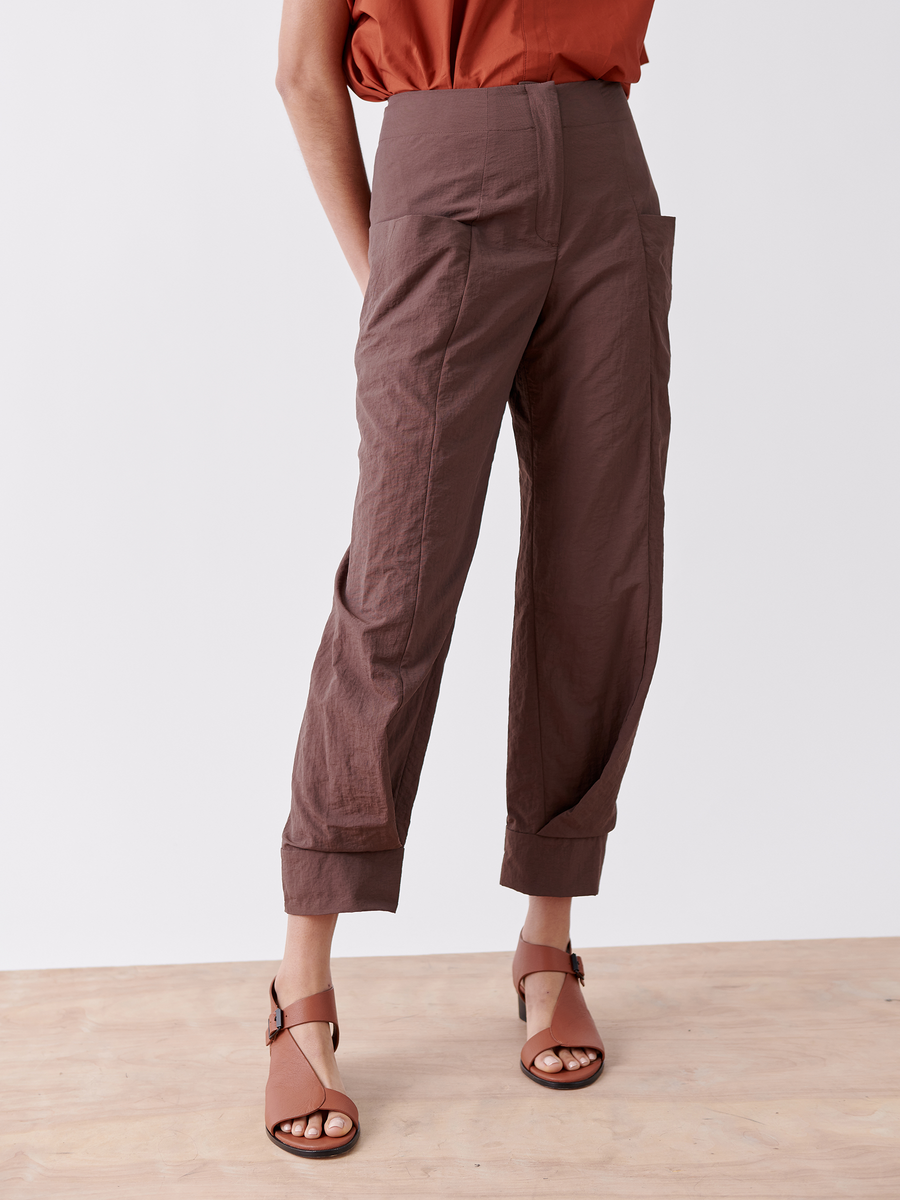 A person wearing Akeo Jodhpur pants from Zero + Maria Cornejo, crafted from lightweight fabric with pockets and a high waist in brown, stands on a wooden floor. They pair the pants with a burnt orange shirt and brown open-toed sandals against a plain white background.