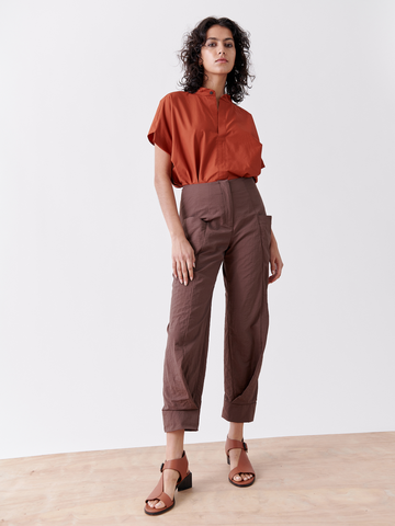 A woman with curly hair confidently stands on a wooden floor against a plain white background, wearing an orange blouse and Zero + Maria Cornejo's Akeo Jodhpur tech taffeta pants in brown, paired with brown sandals.