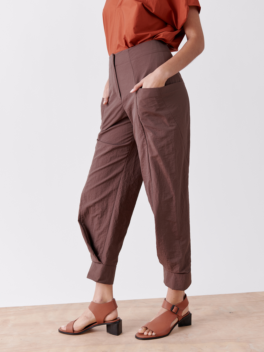 A person wears stylish brown Akeo Jodhpur pants by Zero + Maria Cornejo, featuring a textured water-repellent fabric, and an orange short-sleeve top. Hands in pockets, pants rolled at the ankles, they flaunt brown open-toe heeled sandals, perfect for any weather.