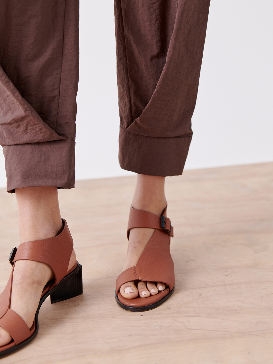 A person is wearing the Akeo Jodhpur by Zero + Maria Cornejo, crafted from lightweight tech taffeta in brown, paired with open-toe sandals featuring ankle straps and buckles, standing on a wooden floor.