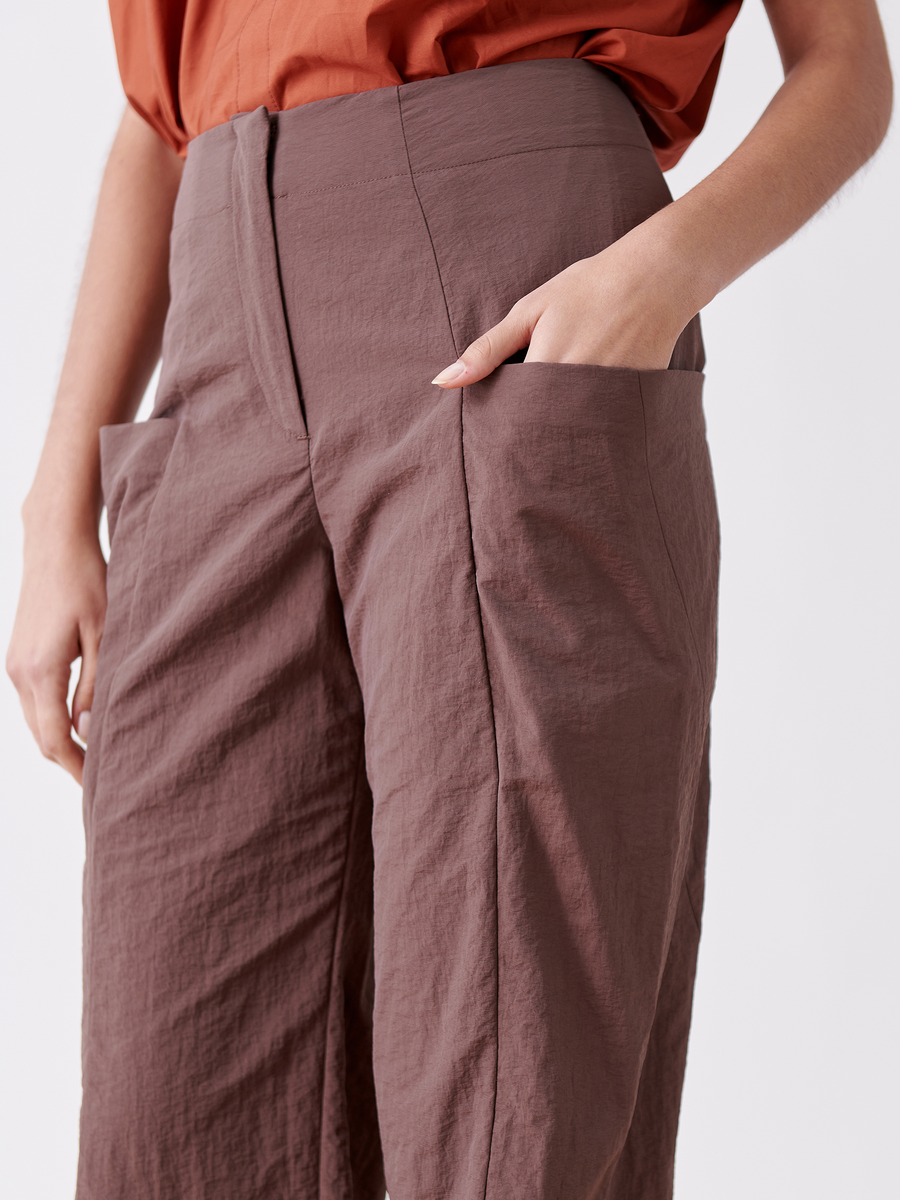 A person in a rust-colored top and Zero + Maria Cornejo's Akeo Jodhpur - Final Sale pants stands with hands in pockets against a white background. These high-waisted pants combine style and practicality with their textured, water-repellent Tech taffeta fabric.