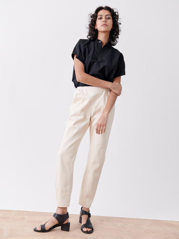 A person with curly hair stands confidently against a plain background, wearing a black shirt and beige Gabi Trousers from Zero + Maria Cornejo. Their relaxed pose is complemented by black heeled sandals and one hand lightly touching the opposite shoulder.