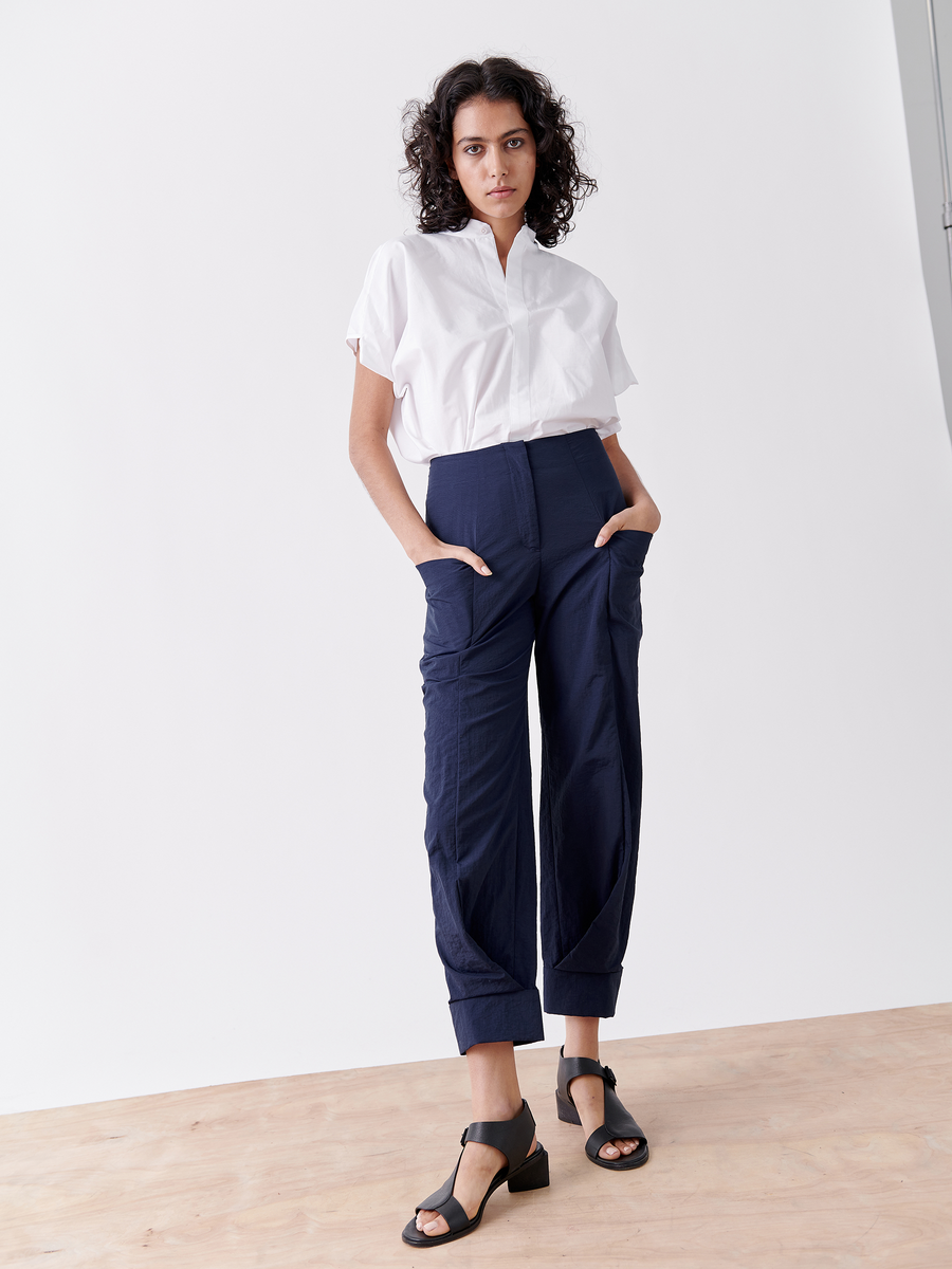 A confident person with curly dark hair stands on a light wood floor against a white background, wearing a white short-sleeved shirt, navy blue tech taffeta pants by Zero + Maria Cornejo, and black sandals. Hands are in pockets of Akeo Jodhpur - Final Sale pants.