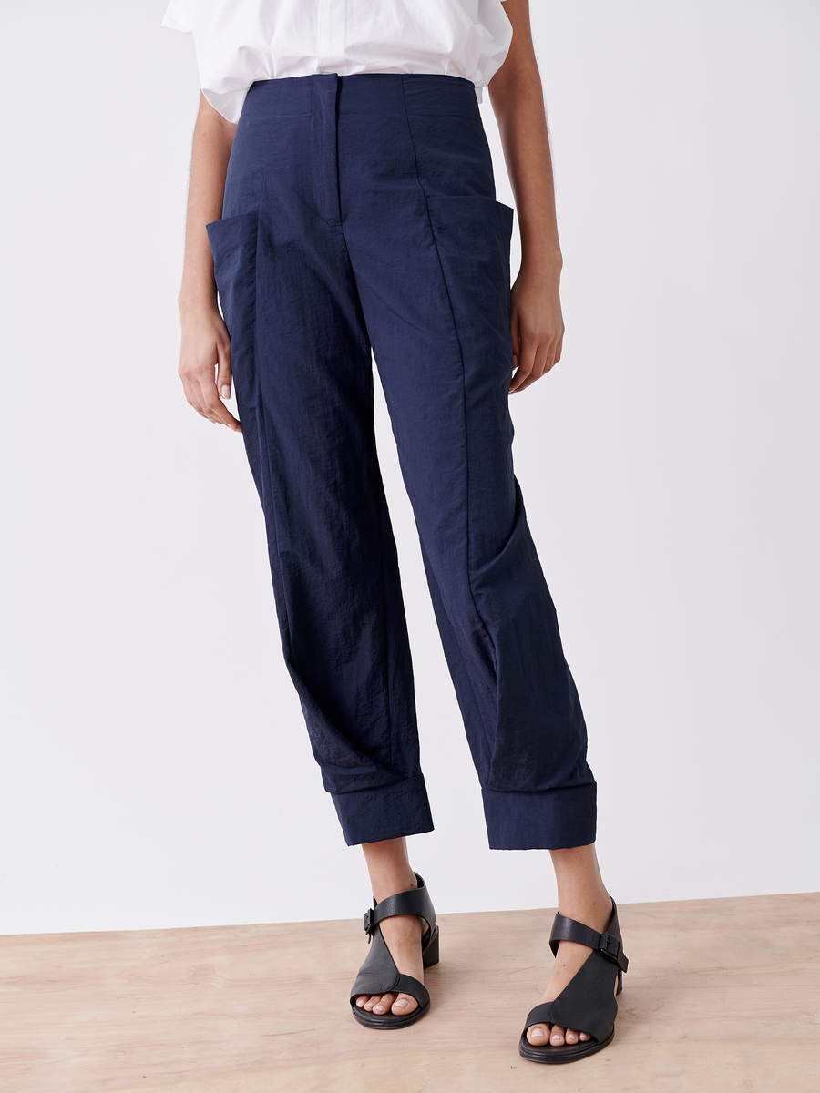 Person wearing Zero + Maria Cornejo's Akeo Jodhpur, denim blue high-waisted tech taffeta pants with large pockets and a slightly tapered fit. Paired with black sandals and a white top, set against a plain white background.