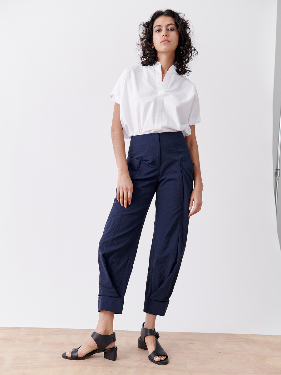 A confident individual with curly hair wears a white collared shirt, navy tech taffeta pants, and dons the Akeo Jodhpur sandals by Zero + Maria Cornejo in black open-toe block heels. The backdrop is minimalistic and light-colored.