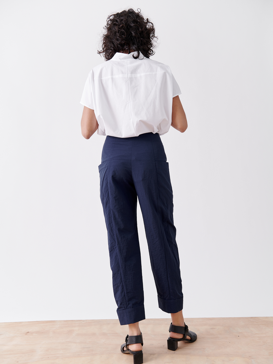 A person with curly hair stands against a white backdrop, wearing a white shirt and dark blue Akeo Jodhpur pants by Zero + Maria Cornejo. The lightweight fabric adds elegance, perfectly paired with black sandals on a light-colored wooden floor.