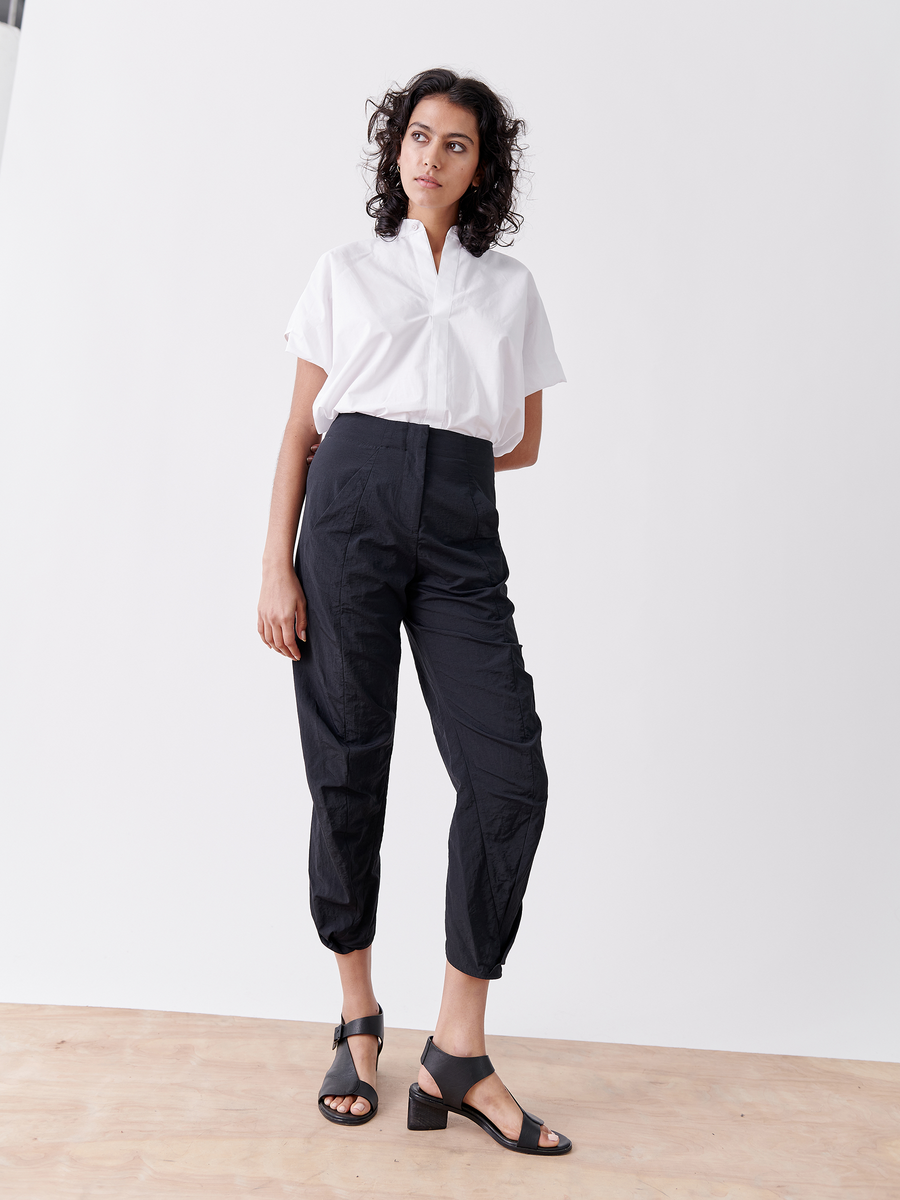 Sporting a white short-sleeved shirt and Zero + Maria Cornejo's Takeo Pant, these cropped black tech taffeta pants offer packable weight, ideal for travel. With black heeled sandals and a serene expression, their pose is relaxed against a white backdrop.