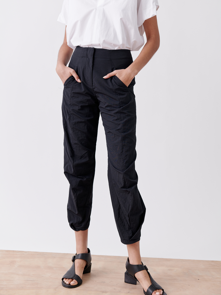 A person in a white short-sleeve shirt and Zero + Maria Cornejo's Takeo Pant - Final Sale, high-waisted tech taffeta pants, stands with hands in pockets. They wear black sandals with a low block heel. The versatile travel ensemble is set against a plain backdrop.