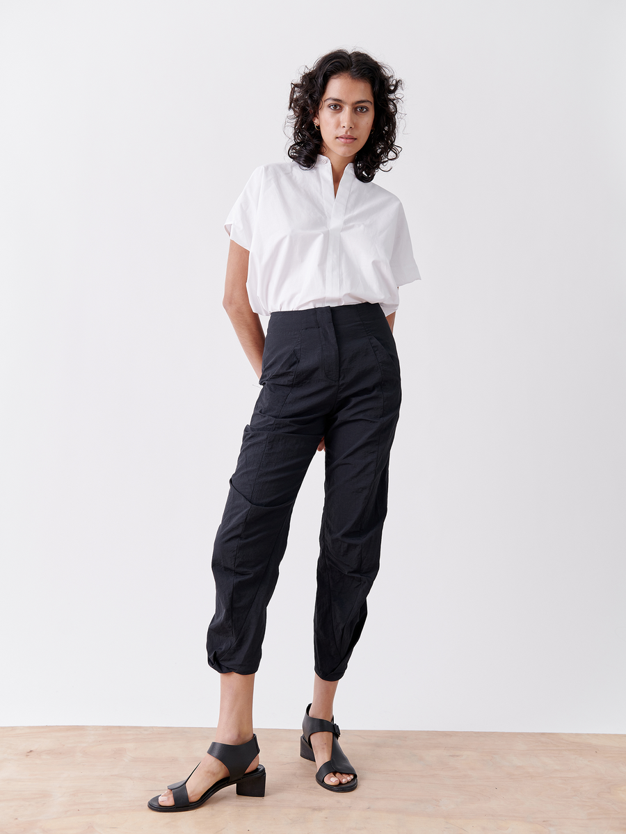 With confidence, a person with curly hair sports a white shirt tucked into Zero + Maria Cornejo's Takeo Pant - Final Sale. These high-waisted black tech taffeta pants are packable for travel. Black low-heel sandals complete the minimalist look against a white background.