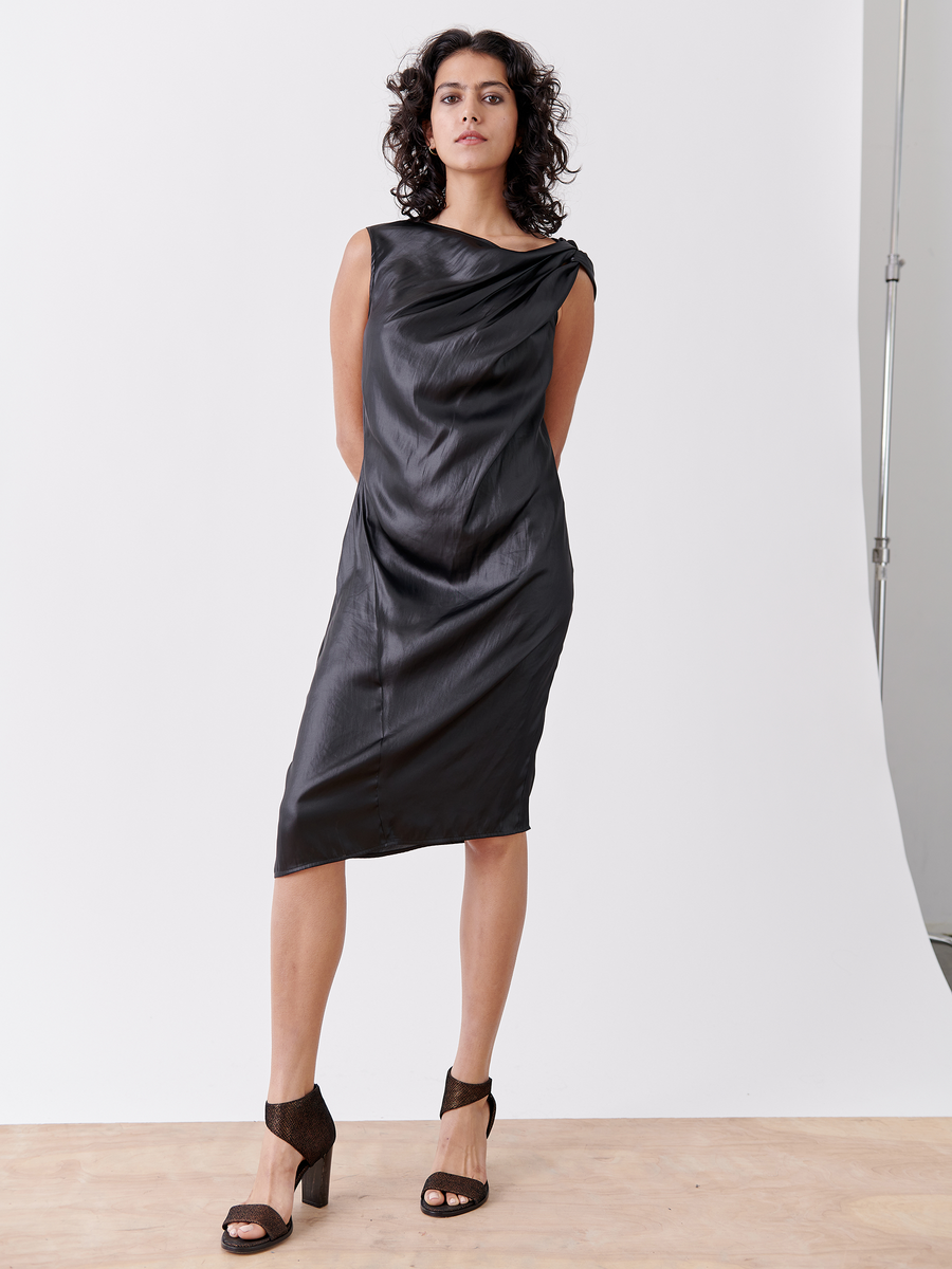 A woman with curly hair stands facing forward in the Ampe Ibit Dress by Zero + Maria Cornejo. The sleeveless, knee-length black cocktail dress showcases an asymmetric knot at the draped neckline. She holds her arms elegantly behind her back, pairing it with black open-toe heels against a white studio backdrop.