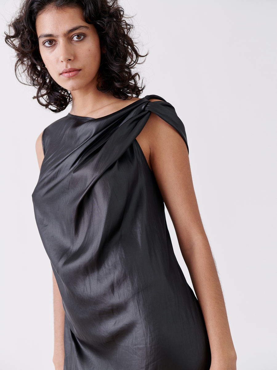 Against a plain light background, a curly-haired person wears the sleek Ampe Ibit Dress - Final Sale by Zero + Maria Cornejo. Made from recycled polyester, it features an asymmetric knot and draped shoulder detail. They gaze forward with a neutral expression in this elegant black cocktail dress.