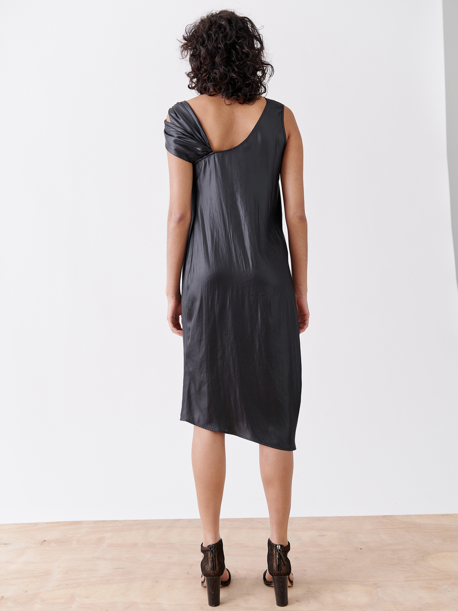 Rear view of a person with curly hair wearing the Ampe Ibit Dress by Zero + Maria Cornejo, a shiny asymmetrical black cocktail dress made from recycled polyester. They stand on a wooden floor against a plain white background, complemented by high-heeled sandals.