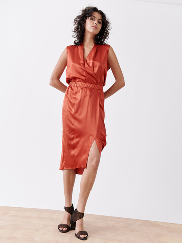 A person with curly hair models the Mio Dress by Zero + Maria Cornejo. This elegant silk charmeuse outfit features a sleeveless design and a knee-high slit. They stand on a light wood floor against a white wall, wearing black open-toe heeled shoes.