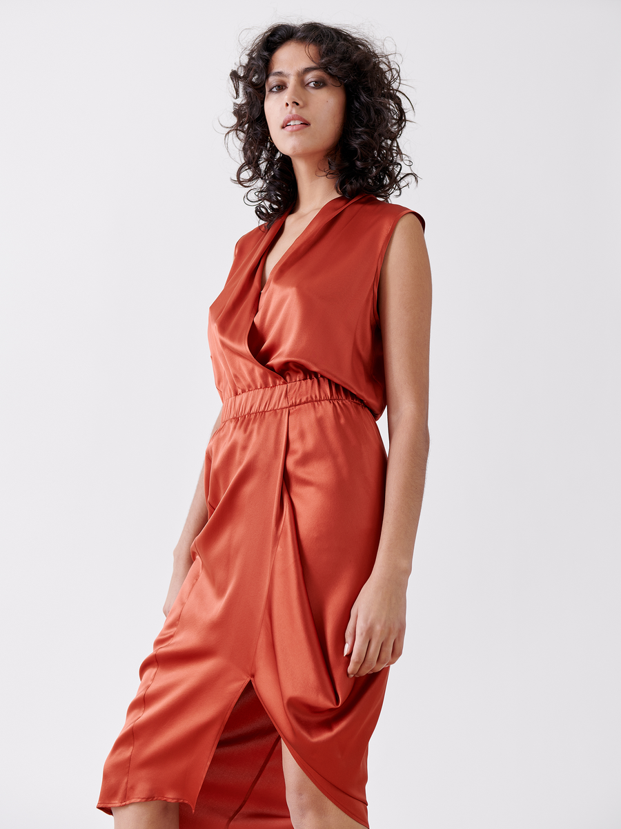 A woman with curly hair wearing Zero + Maria Cornejo's Mio Dress - Final Sale in rust color stands confidently against a plain background. The sleeveless silk charmeuse dress has asymmetrical detailing, a wrap design, a gathered waist, and falls below the knee.