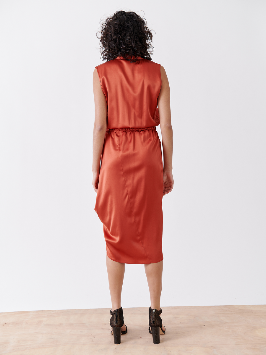 A woman with curly hair stands on a wooden floor, facing away from the camera against a plain white background, wearing the Mio Dress - Final Sale by Zero + Maria Cornejo in orange silk charmeuse and brown ankle boots.