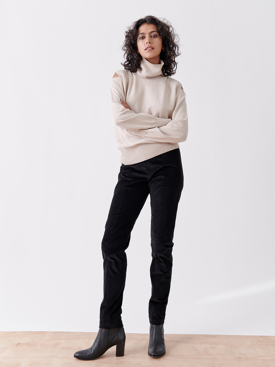 A person with curly hair stands confidently with arms crossed, wearing a beige turtleneck sweater with shoulder cut-outs, Slim Tala Trouser - Final Sale from Zero + Maria Cornejo, and black heeled boots against a plain white background.