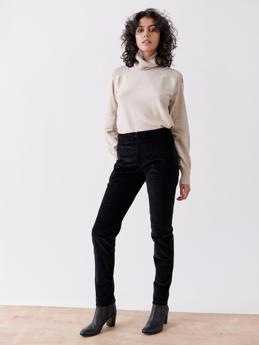 A woman with curly hair stands against a white background wearing a beige turtleneck sweater, Slim Tala Trousers by Zero + Maria Cornejo, and black ankle boots. The trousers' elastic waistband provides comfort and style as she maintains a relaxed posture with her hands by her sides.