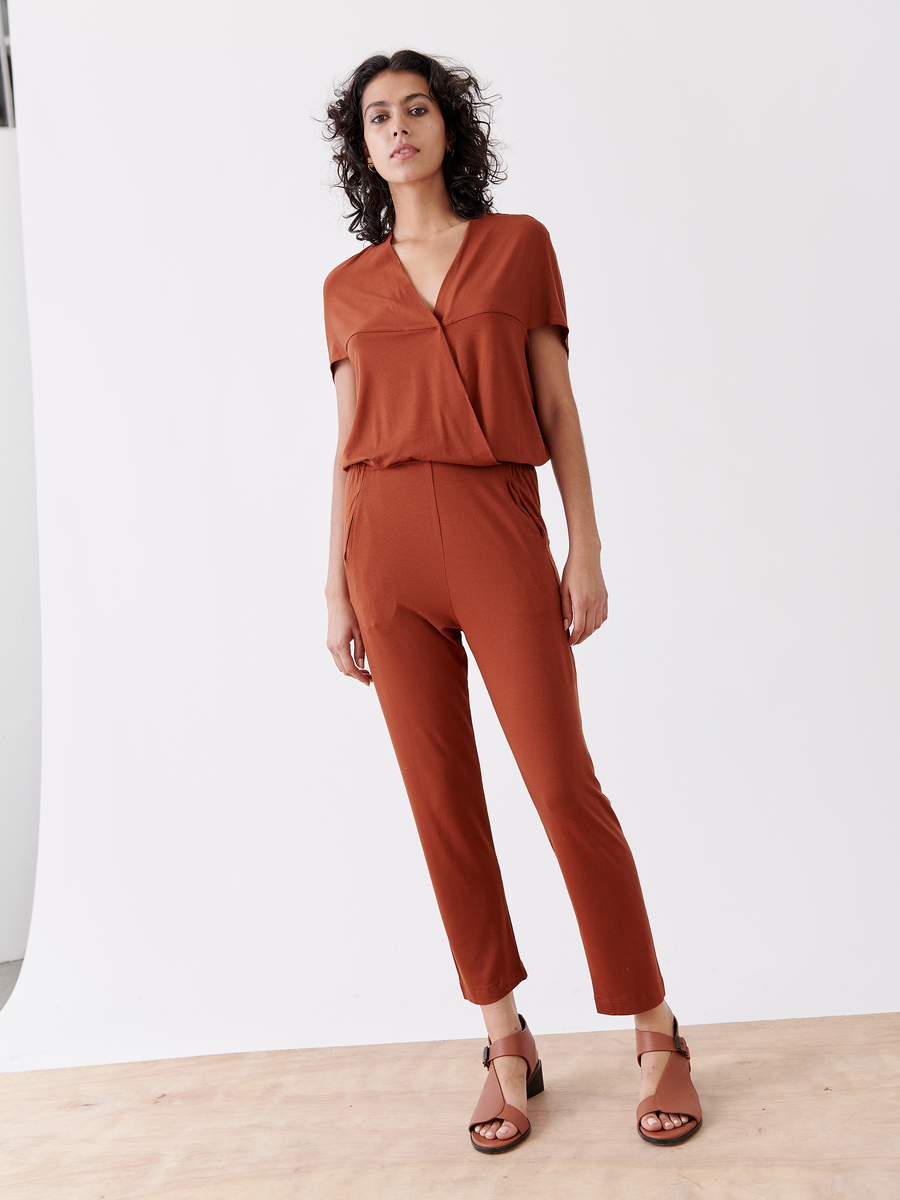 A person stands against a white backdrop in the rust Hex Jumpsuit by Zero + Maria Cornejo, featuring a V-neck, short sleeves, and an elastic waist. They have curly hair and wear brown open-toed shoes.