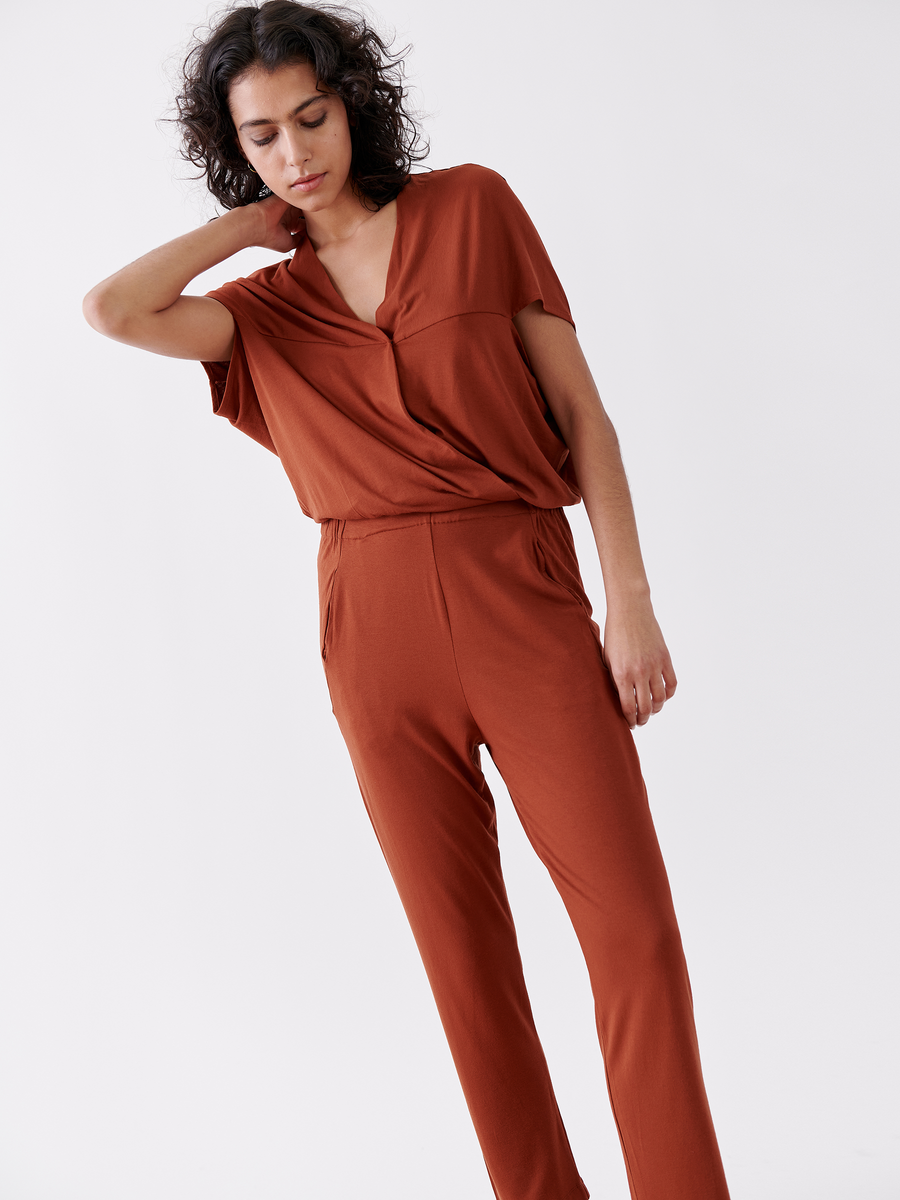 A person wearing the rust-colored Hex Jumpsuit by Zero + Maria Cornejo poses elegantly against a neutral background. One hand touches their neck while the other rests by their side, exuding a calm and relaxed expression.