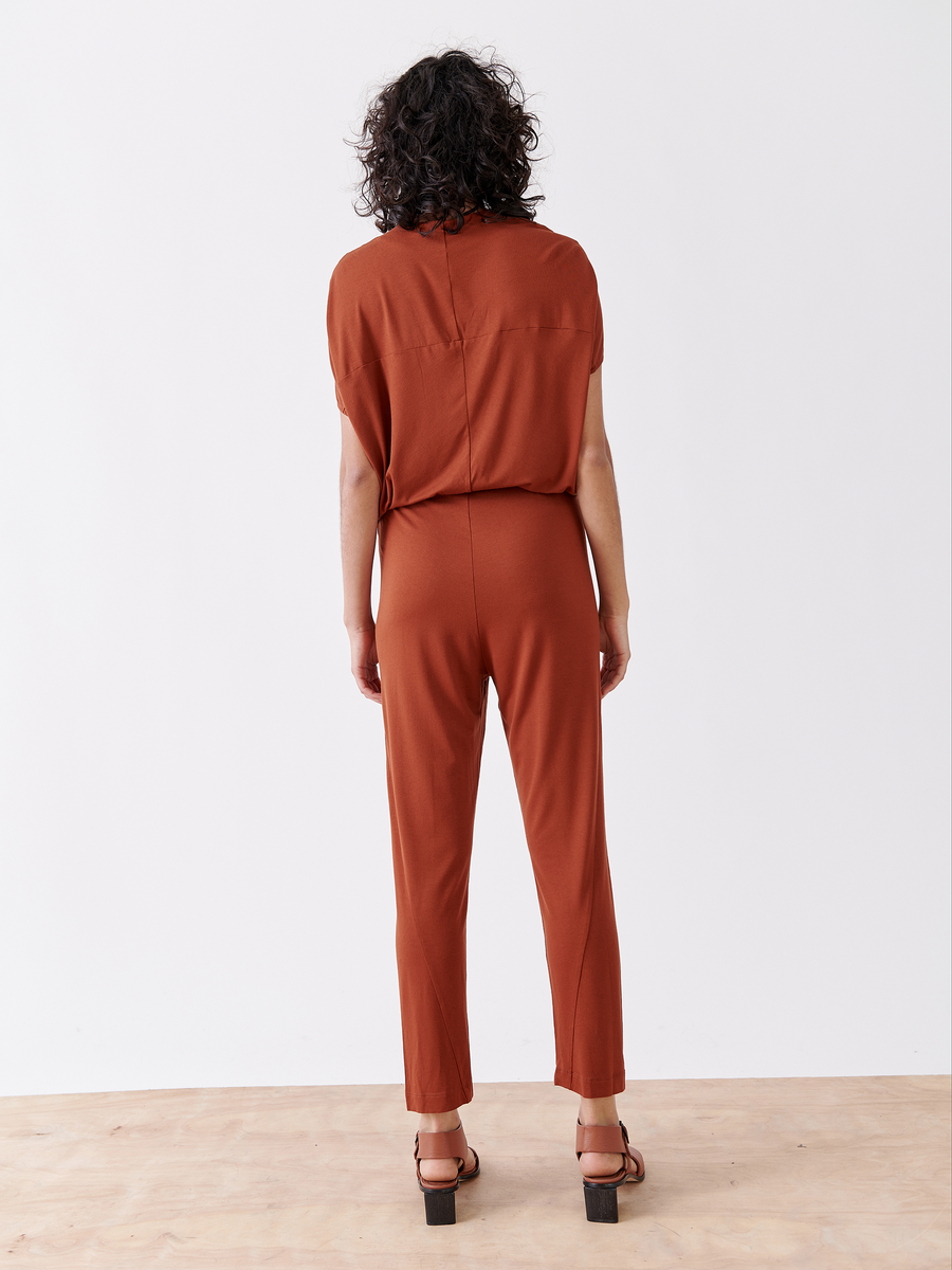 A person with curly hair is shown from the back wearing the rust-colored Hex Jumpsuit by Zero + Maria Cornejo, featuring a v-neck and elastic waist, paired with brown platform sandals. They stand on a light wood floor against a plain white background.