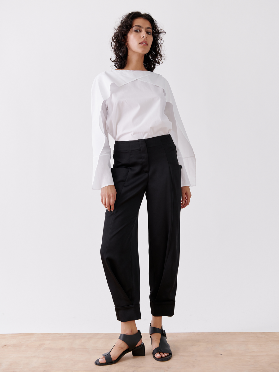 In a minimalist white setting, a person confidently wears the Akeo Jodhpur by Zero + Maria Cornejo—elegant black drape pants made from 100% recycled fabric. Their curly hair matches their black open-toed sandals on the wooden floor, showcasing effortless style designed in New York.