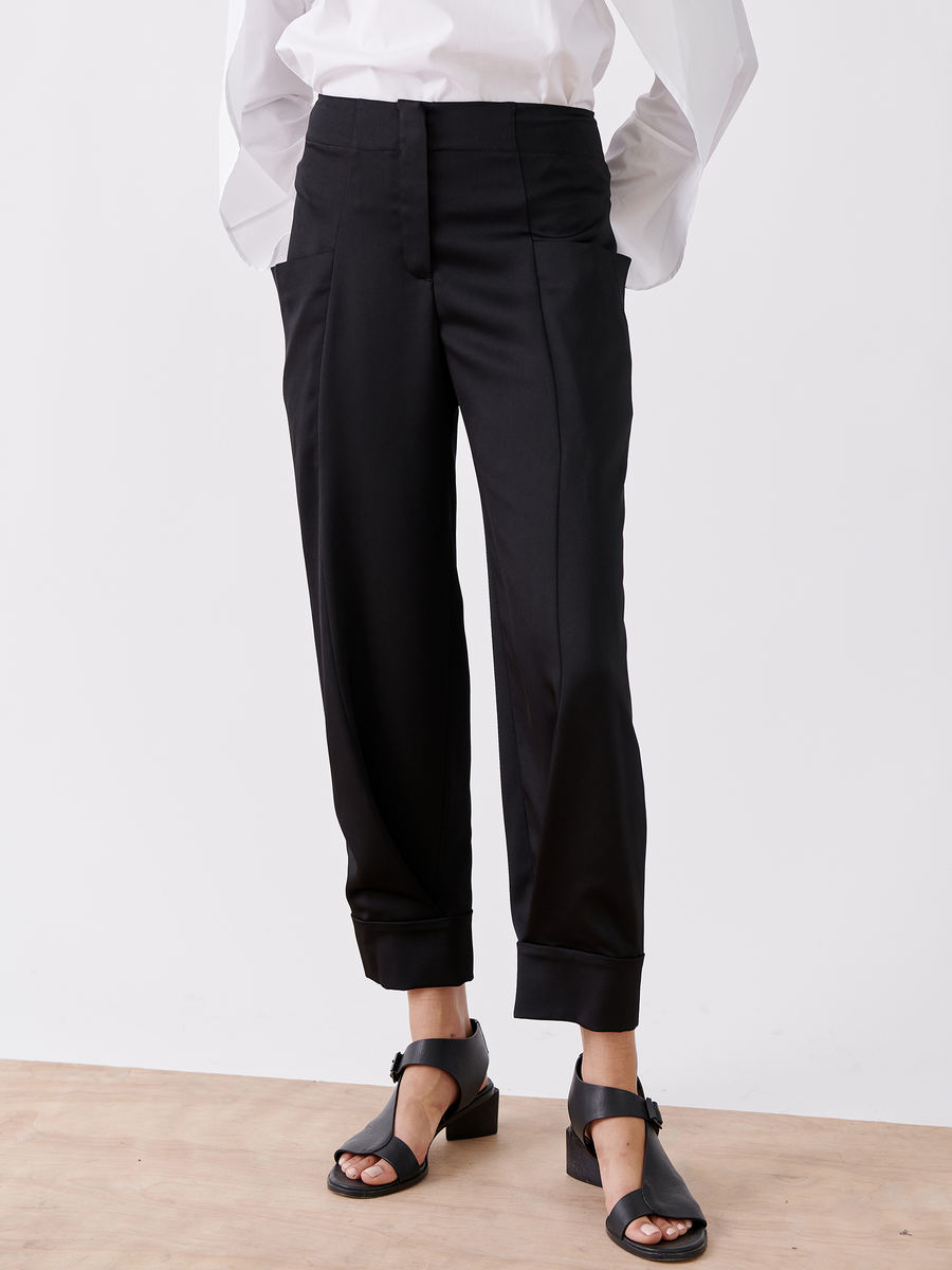 A person wears Zero + Maria Cornejo's Akeo Jodhpur, black recycled drape pants with a high-waisted, cuffed design and side pockets. Paired with a white shirt and black open-toed chunky heel sandals, the outfit showcases sustainable fashion against a plain white wall backdrop.