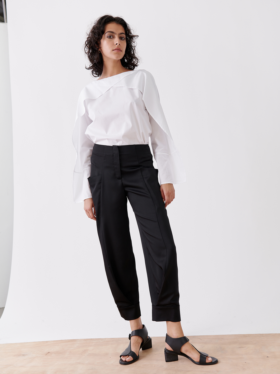 A person with curly hair, standing on a wooden floor against a white background, confidently poses in the Akeo Jodhpur by Zero + Maria Cornejo: a white long-sleeve blouse, black high-waisted recycled drape pants, and black heeled sandals. One knee is slightly bent.