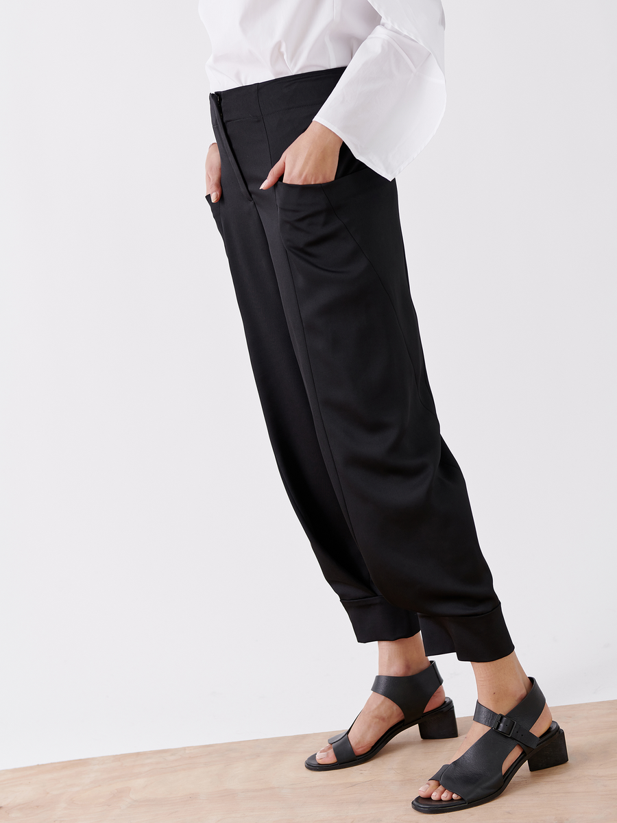 Against a wooden surface, a person wears the Akeo Jodhpur from Zero + Maria Cornejo. The black recycled drape harem pants with side pockets are paired with a white blouse and sleek black heeled sandals, embodying sustainable chic against a plain white backdrop.