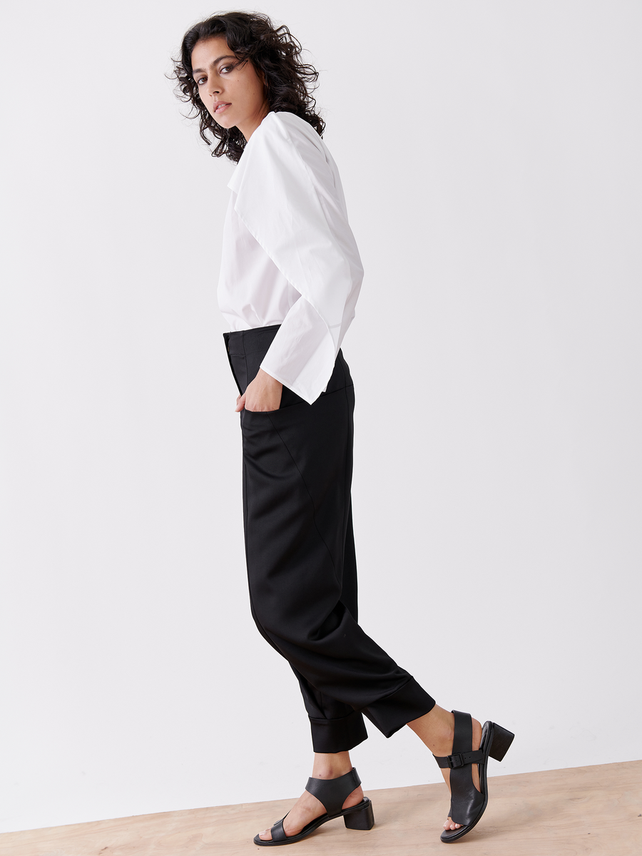 In a minimalist setting, a person with curly hair wears the Akeo Jodhpur by Zero + Maria Cornejo—white long-sleeve shirt and black recycled drape pants made from 100% recycled fabric—paired with black heeled sandals, looking at the camera neutral, embodying sustainable fashion.